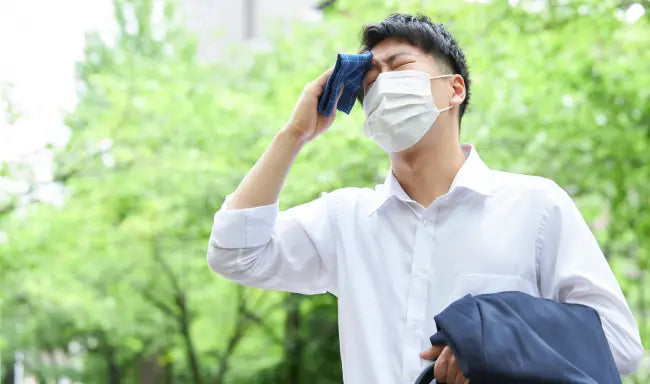 Heat stress awareness training showing an office worker wearing a face mask wiping sweat from his forehead outdoors during hot weather.
