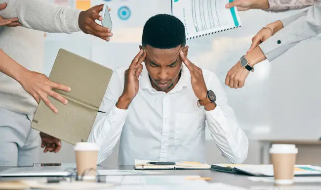 Overwhelmed employee managing workplace stress while colleagues present tasks and business documents in a busy office